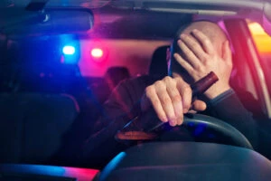 A driver holds his head in one hand and a beer bottle in the other while behind the wheel of his vehicle. To report a drunk driver in New York, call 911 immediately and give the dispatcher key details to help law enforcement find the driver.