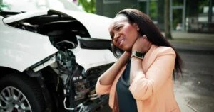 A woman holds her neck in pain next to a wrecked vehicle and wonders when is it too late to go to the doctor after a car accident.