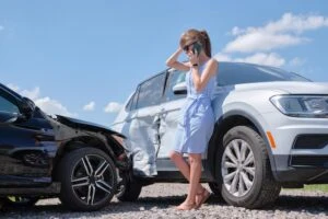 A woman in a blue dress stands by her white SUV and a black car that T-boned it. She talks on her cell phone and holds her head, wondering what to do after a car accident in Las Vegas. 