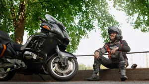 Motorcyclist sitting on a curb wearing full protective motorcycle attire with his arms resting on his knees and smile on his face.