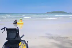 An empty wheelchair on a beach with a child playing in the water by an inflatable duck ring