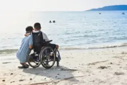 A child with cerebral palsy sits in a wheelchair next to his parent looking out at the ocean.