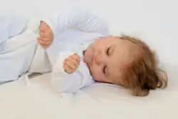 an infant in a white pajama sleeper is lying down and holding one arm in an awkward position