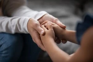 Close up compassionate young foster parent holding hands of little kid girl, giving psychological help, supporting at home.