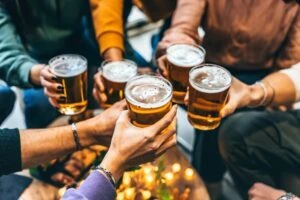 Five hands clink beer glasses together while somebody wonders what typical frat hazing is.