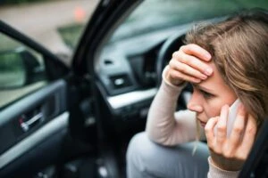 A distressed woman sits in the driver’s seat of her car with her door open after an accident. She holds her head and her phone to her ear, asking, “What should I do if I am hit by a commercial vehicle in Dayton?”