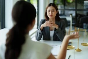A lawyer sits at a table with her client, a woman with dark hair, who asks, “What legal protections exist for hazing victims?”