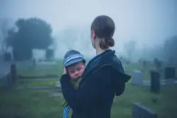 A young woman is holding a baby and standing in a graveyard.