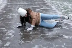 A young woman wearing a hat, gloves, and jacket is lying on an icy walkway.