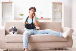 An injured man with an arm sling and foot cast sitting on a couch with his helmet, calling a motorcycle accident lawyer.
