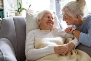 A young blonde woman smiles at an elderly woman in an armchair who is wondering about the difference between long term disability insurance and long term care insurance.