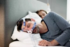 A man who meets the VA rating for sleep apnea sleeps in bed with a CPAP machine with his wife sleeping beside him.