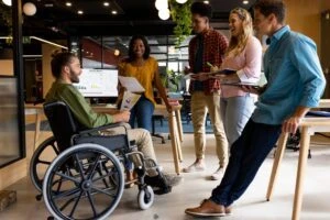 Work colleagues, including an employee on disability, talk during a casual office meeting. If you have a disability and want to know whether you will lose your benefits if you work part time, our disability lawyer can clarify how working part-time might affect your benefits.