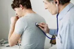 A young man coughs as an older male doctor holds a stethoscope to his back.