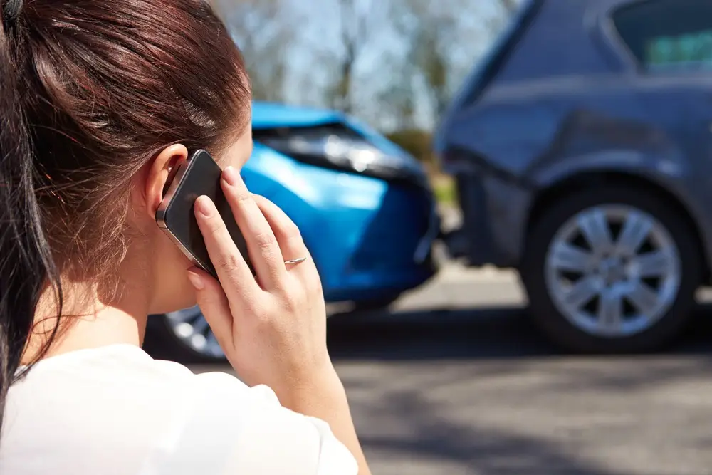 A woman viewed from behind is making a phone call on a cell phone. In the background, two cars can be seen that have recently been involved in a rear-end collision. The woman can get medical treatment and pre-settlement funding to pay bills from Rockpoint Legal Funding.