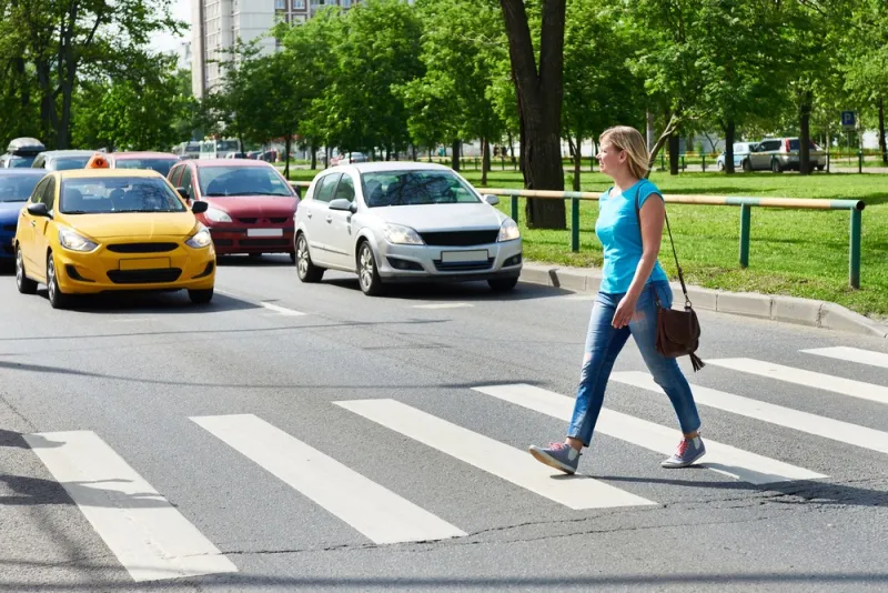 A woman, in jeans and a t-shirt is seen walking across a street in a crosswalk on a bright, sunny day. A green park can be seen behind her. Rockpoint Legal Funding provides cash advances for plaintiffs in personal injury lawsuits who have been struck by a motor vehicle.