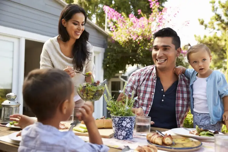 A family enjoying an outdoor meal on a sunny day. Man, woman and two small children are around a picnic table and the woman is standing and serving salad from a large bowl. The woman and man are smiling. Rockpoint Legal Funding, the best lawsuit loan company.