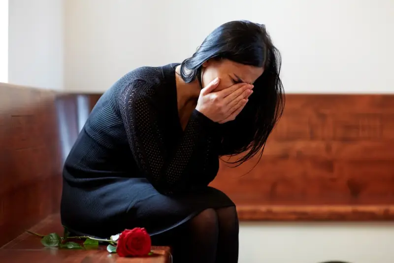 A dark-haired woman sits on a wooden bench. There is a single long-stem red rose next to her. She is wearing a black dress and her pained face is in her hands as if she is crying.