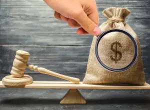 A gavel sits on one end of a plank while a bag of money magnified with a magnifying glass sits on the other end.