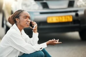 A woman sits on the ground in front of her car and makes a phone call to ask, “What should I do if I’m an immigrant in a car accident?”
