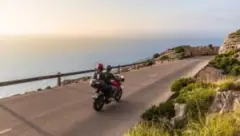 A motorcyclist and their passenger drive by the ocean.