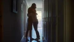 Two grieving people hug in a dimly lit hallway.