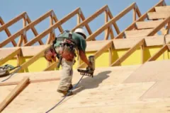 A construction worker safely climbs a roof while using a power tool.
