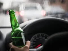 An image of a driver’s hands on a steering wheel while holding an open beer bottle.