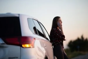 Woman leans against car and uses cell phone to report minor car accident to the insurance company.