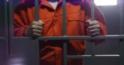 Elderly prisoner in orange uniform holds metal bars, stands in prison cell.