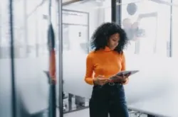 Professional Black businesswoman using tablet in modern office setting, focused and serious, wearing casual orange sweater and jeans, exemplifying mobile business management.