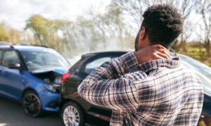 A young man in a plaid shirt has his hand on his neck and is standing near a car accident scene.