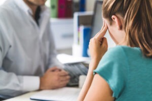 A young woman is seated across from a doctor and has two fingers on her temples.
