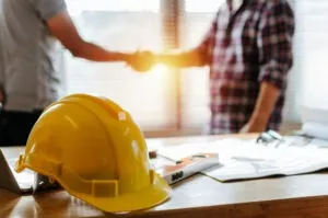 An architect and a construction worker shake hands backlit by sunlight. A construction hat and site plans are on a desk in the foreground.