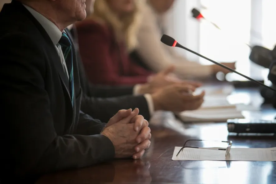 Man in a suit, hands folded, speaking into a microphone before a Texas licensing board. Professional license defense attorneys in the background.