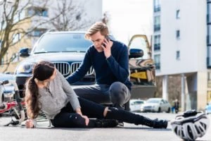 Injured bicyclist lying on the road in front of a car. The driver comforts the bicyclist while making a call on their cellphone