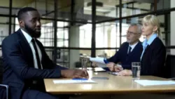 A black business professional sits at a desk with a white male and white female, asking how to prove racial discrimination.