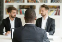 Rear view of a black businessman at a desk in front of two white male coworkers sitting on the opposite side, showing an example of racial discrimination at work.