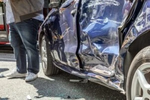 Crashed blue car with victim standing next to door wondering what happens if you are in an accident in an Uber