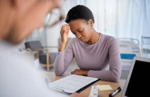 A woman stands before a receptionist and holds her head, looking down at her hospital bill with concern and wondering if health insurance covers car accidents in Florida.