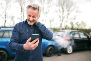 A man stands by two cars damaged in a rear-end accident. He holds his neck and looks at his phone, researching the cost of a car accident lawyer.