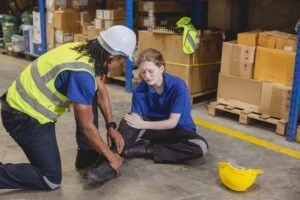 A young woman warehouse employee sits on the ground and holds her leg in pain as a coworker tries to help her up. She wonders how long slip and fall settlements take.