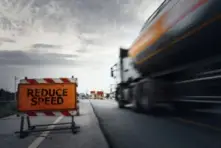 A semi-truck speeding past a sign on the freeway reading REDUCE SPEED