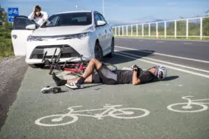 An injured male bicycle rider is lying on the ground in the bike lane in front of a white car.