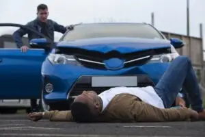 A man is lying in front of a blue car after a pedestrian accident.