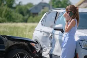 A woman leans against a damaged car and talks to an insurer on the phone about total loss value.