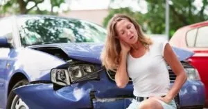 Woman wearing a white t-shirt and jean shorts who has been injured in an accident while driving for Postmates sitting in front of a blue sedan.