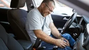 Man in a t-shirt and jeans sitting in the driver's seat of a car who has both hands on his right knee after being injured in an accident while driving for Uber.