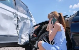 Woman in a blue dress kneeling down next to a black sedan looking at a white SUV in front of her and speaking with someone on her smartphone to report vehicle damage after a DoorDash car accident