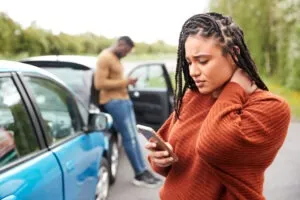 Woman looking down at her cell phone with two cars to her right and a man leaning against one of the cars, with the woman trying to determine who is liable for a Grubhub car accident involving these vehicles